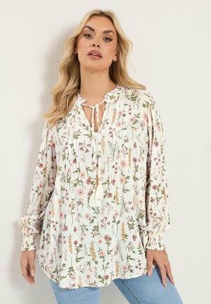 Pitkähiuksinen nainen, jolla on pitkähihainen valkoinen kukallinen paita tupsusolmujen kanssa ja vaaleansiniset farkut, yksiväristä taustaa vasten.