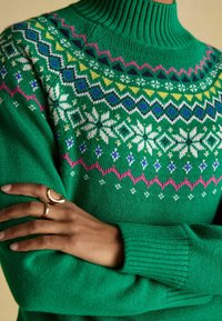 Grüner Strickpullover mit hohem Rippkragen, geometrischen Mustern in Weiß, Blau, Pink und Gelb rund um den Halsausschnitt und den Körper.