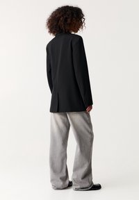 Blazer noir au design cintré, fente unique à l'arrière, associé à un jean large gris clair et des chaussures noires.