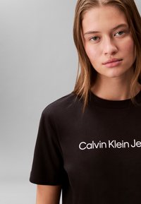 Czarny bawełniany t-shirt z krótkimi rękawami, z białym logo „Calvin Klein Jeans” na piersi, gładką fakturą i klasycznym krojem.