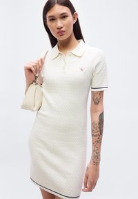 Λευκό πλεκτό φόρεμα πόλο με κοντά μανίκια, με μικρό λογότυπο, υφασμάτινο μοτίβο και μαύρη λεπτομέρεια στο τελείωμα. Κρατάει μια μπεζ τσάντα.