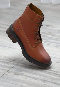 Bota de tobillo de cuero marrón con diseño de cordones, superficie texturizada y suela de goma. Presenta un logotipo en el lateral y detalles de costura.
