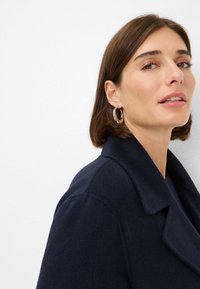 Manteau en laine navy avec col cranté et texture douce. Le modèle porte de grandes boucles d'oreilles argentées, mettant en valeur un profil en gros plan.