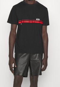 Camiseta de algodón negra con una franja horizontal roja y el logo blanco "GCDS" en el pecho, combinada con pantalones cortos de cuero negros.