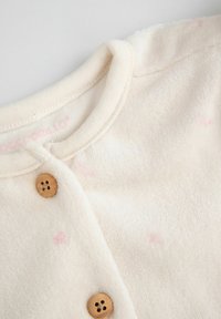 Miękka, kremowa odzież niemowlęca z okrągłym dekoltem, dwoma drewnianymi guzikami i delikatnymi różowymi kropkami na materiale.