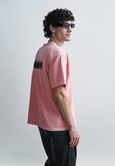 Giovane con capelli ricci che indossa occhiali da sole neri, una maglietta rosa con testo sbiadito sul retro e pantaloni neri, in piedi di lato.