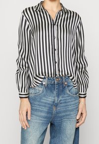 Kvinna klädd i en svart och vit vertikalt randig långärmad skjorta instoppar i högmidjade, blekta blå jeans, med ett armband på höger handled.