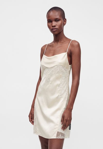 Femme portant une robe en satin crème sans manches avec des panneaux en dentelle et des fines bretelles, sur un fond clair uni.