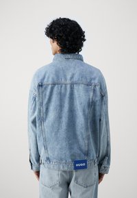 Giacca di jeans blu chiaro con colletto, maniche lunghe e taglio dritto. Presenta un'etichetta blu con "HUGO" sull'orlo posteriore. Senza segni di usura visibili.