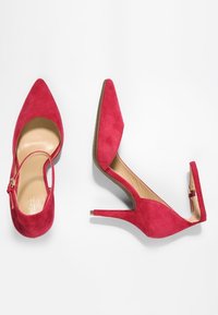Pump rosse in suede con tacchi alti, punta affusolata e retro aperto. Presentano una sottile striscia alla caviglia e un elegante tacco a spillo. Accenti in hardware dorato.