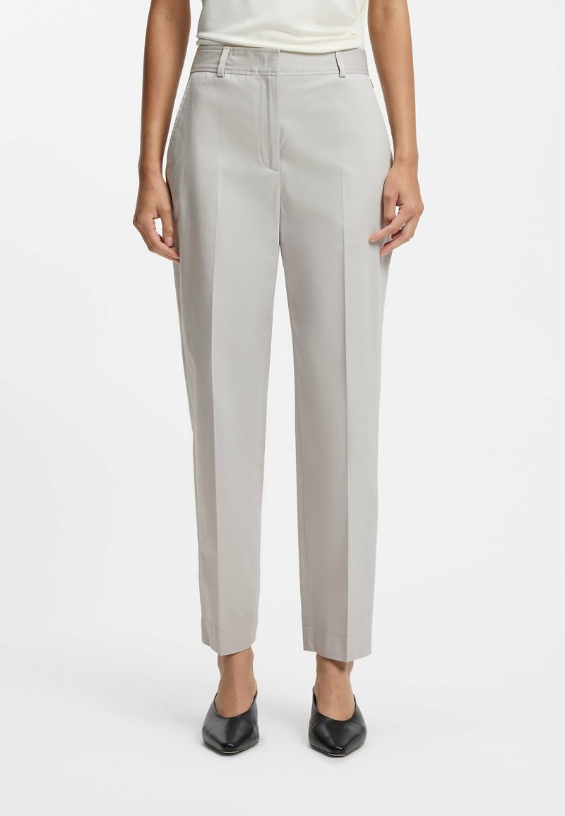 Femme portant un pantalon gris clair tailleur avec plis devant, associé à des chaussures noires à bout pointu, debout sur un fond uni.