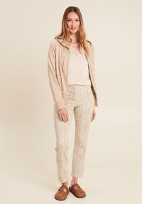 Helle beigefarbene Zip-Hoodie, über einem pinken Oberteil und cremefarbenen, mit Spitzenmuster versehenen Hosen getragen. Braune Slipper runden das Outfit ab.