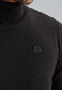 Maglione nero a collo alto con una texture liscia, presenta un piccolo etichetta nera sul petto. Collo a coste e dettagli di cuciture visibili.