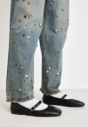 Jambes vêtues d’un jean denim bleu clair avec des clous métalliques et des ourlets effilochés, des chaussettes blanches et des ballerines Mary Jane en cuir noir avec une bride à boucle.