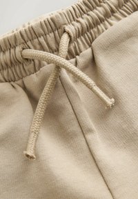 Béžové tepláky mají elastický pas s tkaným stahovacím šňůrkem, jsou vyrobeny z měkké, texturované látky a mají volný střih.