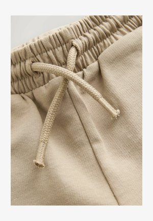 Bež trenirka ima elastični pojas s tkanim vezicama, izrađena je od mekanog, teksturiranog tkanja s opuštenim krojem.