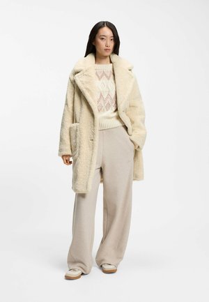 Kvinde iført en cremefarvet tekstureret frakke, mønstret striktrøje, løse beige bukser og lyse sneakers, stående mod en hvid baggrund.