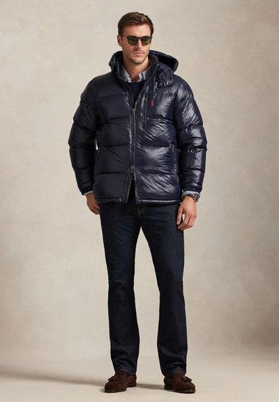 Man wearing black puffer jacket with hood, dark jeans, brown shoes, and sunglasses against plain beige background.