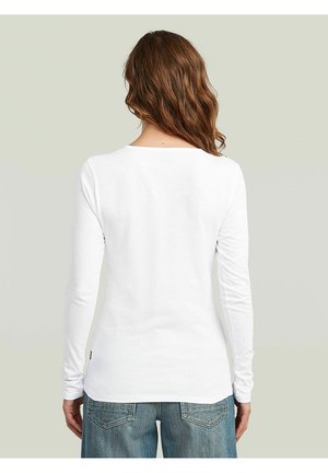 Femme aux cheveux bruns ondulés portant un t-shirt blanc uni à manches longues et un jean bleu, vue de dos sur un fond clair.