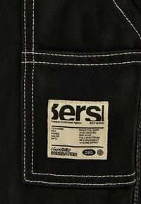 Mustan denim-kankaan, jossa on kontrastoiva valkoinen ommel. Tägissä lukee "bers" ja siinä ovat tuotteen tiedot suorakulmaisella ommellulla tarralla.