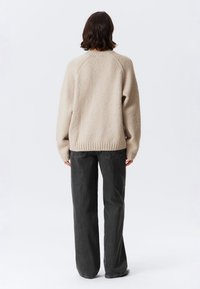 Persoon met schouderlang bruin haar, gekleed in een beige gebreide trui en losse zwarte jeans, staande met de rug naar de camera op een effen witte achtergrond.