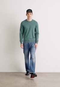 Green sweater with a round neckline and ribbed cuffs, paired with faded blue jeans. Model wears a black cap with "BOSS" logo and black shoes.