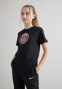 Pusaudze ar gariem pīnēm, tērpusies melnā Paris Saint-Germain T-kreklā un melnās Nike biksēs, ar rokām kabatās, stāv pie baltas sienas.