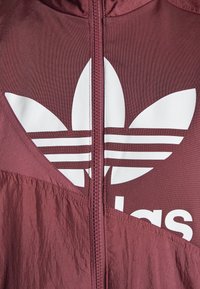 Kaštanově hnědá bunda na zip s lesklou tkaninou, s velkým bílým logem Adidas a třemi vodorovnými pruhy. Texturovaná oblast kolem loga.