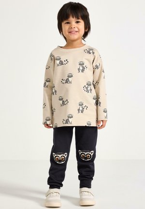 Maglietta beige a maniche lunghe con stampa di lemure, abbinata a pantaloni della tuta blu navy con volti di lemure ricamati e scarpe da ginnastica bianche.