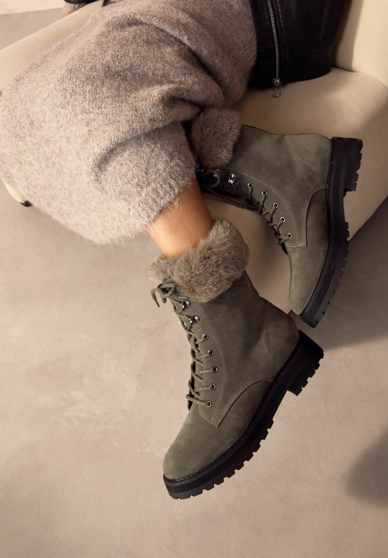 Gröna mocka snörstövlar med faux-fur trim, robust sula och metallhål, placerade på en neutral bakgrund.