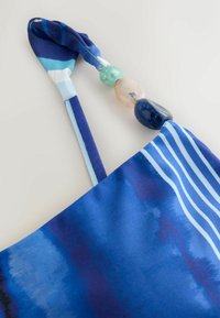 Tissu bleu à motifs avec fines bretelles, comportant trois grandes perles décoratives en vert, pêche et bleu foncé près de l'attache de la bretelle.