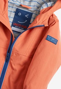 Oranssi tuulenpitävä takki, jossa on sininen vetskari, sininen tilkkupaita, jossa on hymyilevä kasvokuva, ja raidallinen harmaa vuori.