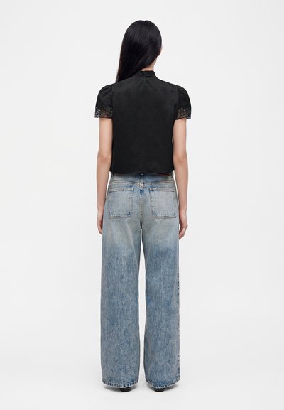 Femme aux longs cheveux noirs portant un haut noir à manches courtes avec des poignets en dentelle et un jean bleu taille haute à jambes larges, debout de dos.
