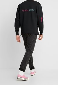 Persona che si allontana indossando jeans neri, una maglietta a maniche lunghe nera con testo multicolore sulla schiena, calze bianche e scarpe da ginnastica rosa.