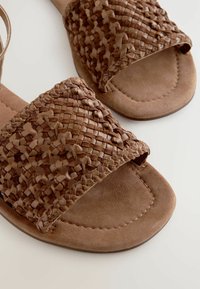 Brune vævede sandaler lavet af tekstureret materiale med en flad sål og en bred rem. Har en glat overflade og et indviklet fletdetalje.