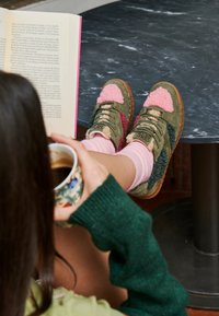 Gröna och rosa texturerade sneakers med mjuka detaljer, matchade med ljusrosa strumpor. En mönstrad mugg och en öppen bok syns i händerna.