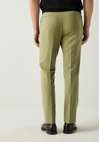 Groene slim-fit broek met twee achterzakken en persplooien. Draagt met zwarte schoenen. Soepele stof met een subtiele glans.