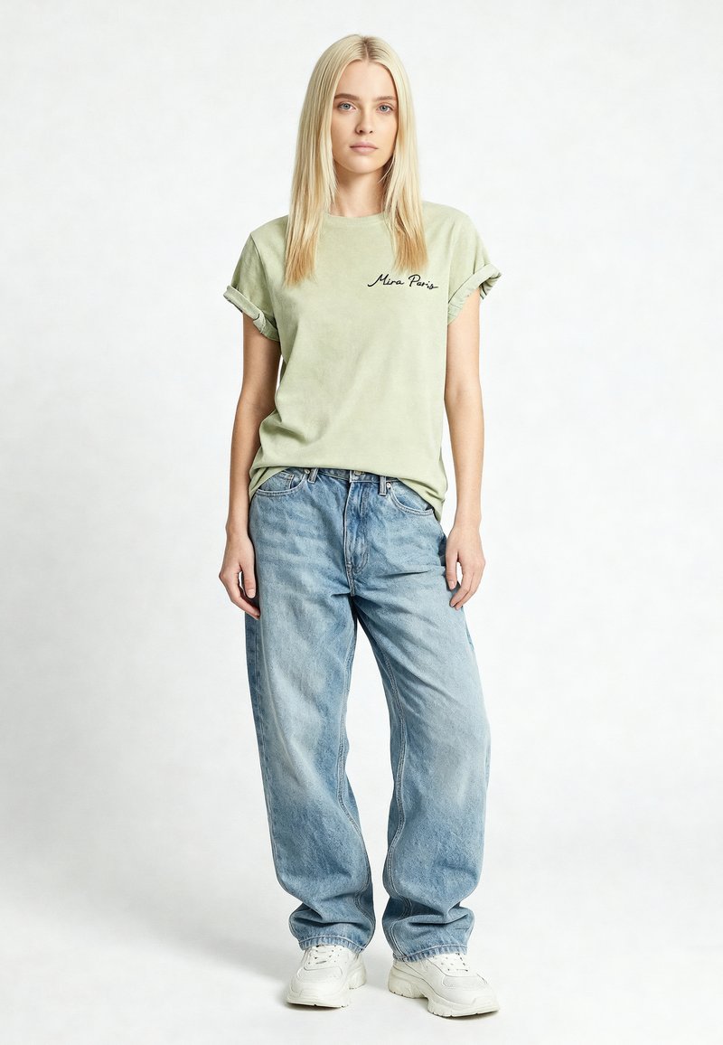 Donna con capelli biondi lisci che indossa una maglietta verde chiaro con maniche arrotolate e jeans azzurri larghi, in piedi davanti a uno sfondo bianco uniforme.