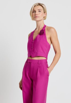 Donna con capelli biondi corti che indossa un gilet smanicato magenta con dettaglio floreale e pantaloni a vita alta abbinati, in piedi con la mano in tasca.