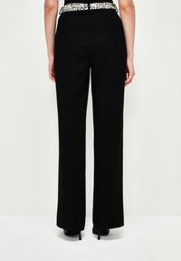 Zwarte wijde broek met een gemêleerde witte en zwarte patroonband. Kenmerkt zich door een achterzak en een gladde stofafwerking.