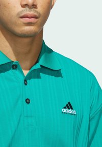 Tyrkysové polo tričko s vertikálními pruhy, límečkem a černými knoflíky. Na levém prsu se nachází černo-bílé logo Adidas.