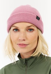 Růžový žebrovaný kulich s ohrnutým lemem a malou černou jmenovkou. Nosený s zelenou zipovou mikinou. Detailní záběr na čepici a obličej, neutrální pozadí.