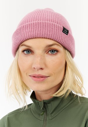 Roze geribbelde beanie met een opgevouwen rand en een klein zwart label. Draagt een groene ritstrui. Close-up van de muts en het gezicht, neutrale achtergrond.