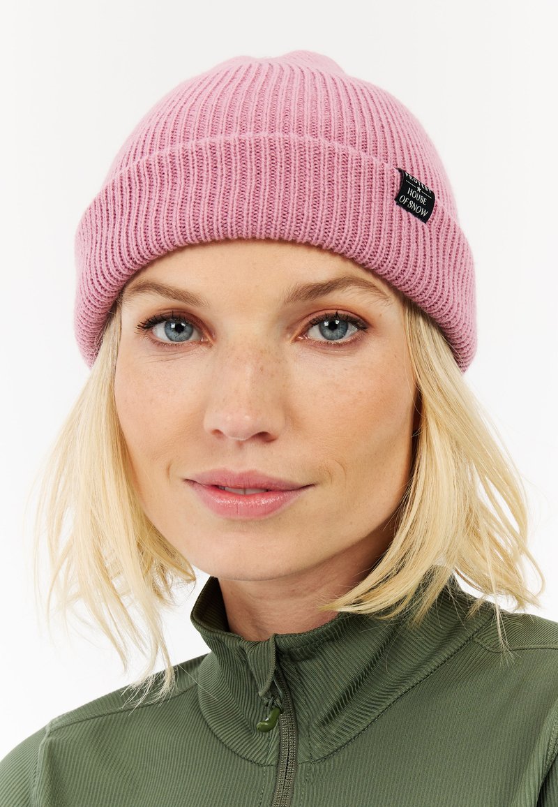 Růžový žebrovaný kulich s ohrnutým lemem a malou černou jmenovkou. Nosený s zelenou zipovou mikinou. Detailní záběr na čepici a obličej, neutrální pozadí.