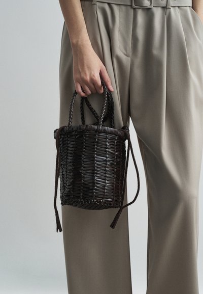 Saco de cesta castanho escuro trançado com duas alças curtas, apresentando uma forma cilíndrica e fecho com cordão ajustável, segurado por uma mão.