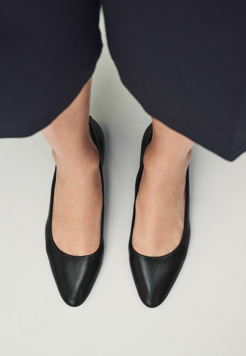 Scarpe basse in pelle nera con una consistenza liscia, design a punta e taglio basso sui lati, su uno sfondo chiaro.