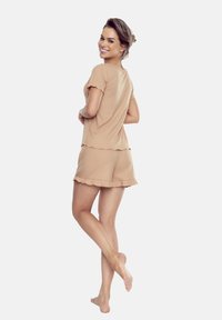 Kobieta w beżowym dwuczęściowym zestawie piżamowym uśmiecha się przez ramię, w krótkich rękawach i falbaniastych brzegach, stojąc boso na jednolitym tle.