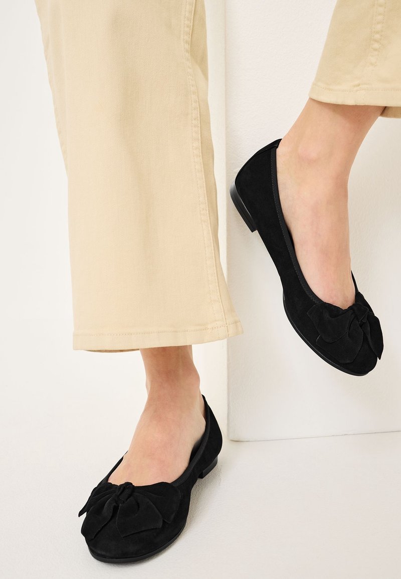 Ballerines en daim noir avec un détail de nœud bien en vue sur le bout, associées à un pantalon court de couleur claire. Texture lisse et talon bas.