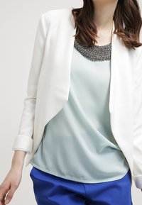 Blazer bianco con un davanti drappeggiato, indossato sopra un top verde chiaro con un collo decorato di perline, abbinato a pantaloni blu. Texture morbide e design moderno.