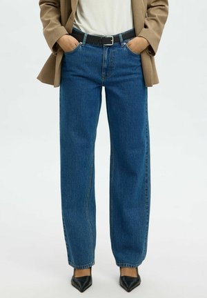 Person trägt gerade geschnittene blaue Jeans, schwarzen Gürtel, beige Blazer, weißes Hemd und schwarze spitze Schuhe, Hände in den Taschen.